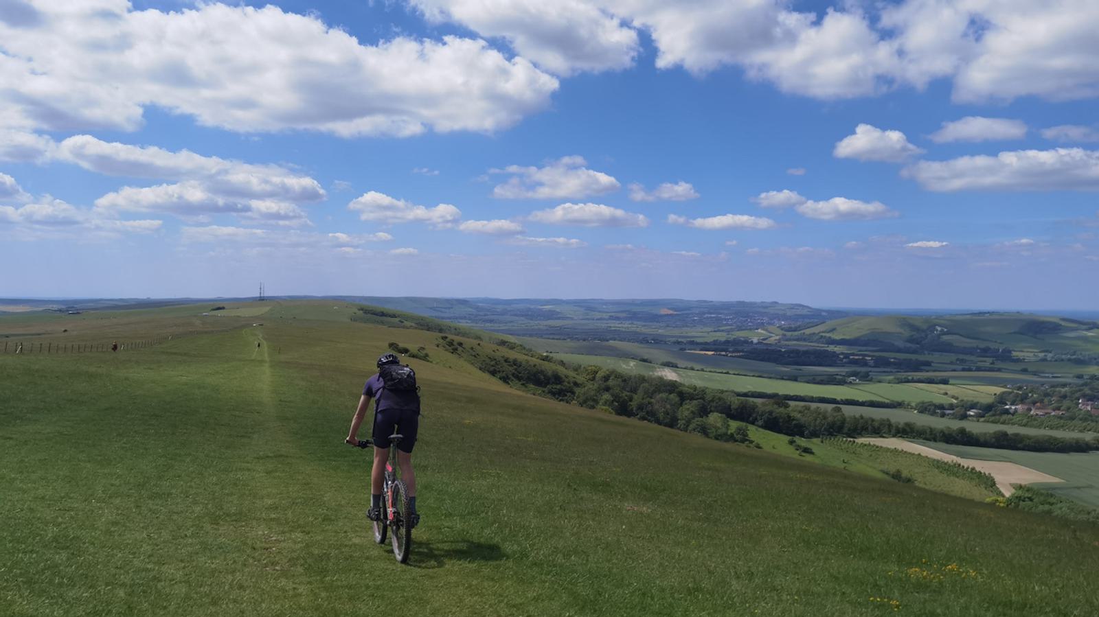 A cyclist rides along a grassy path on a hilltop, surrounded by a vast mountain landscape under a clear, partly cloudy sky. Rolling green hills and distant valleys stretch out below, with rural fields and patches of forest visible in the distance.