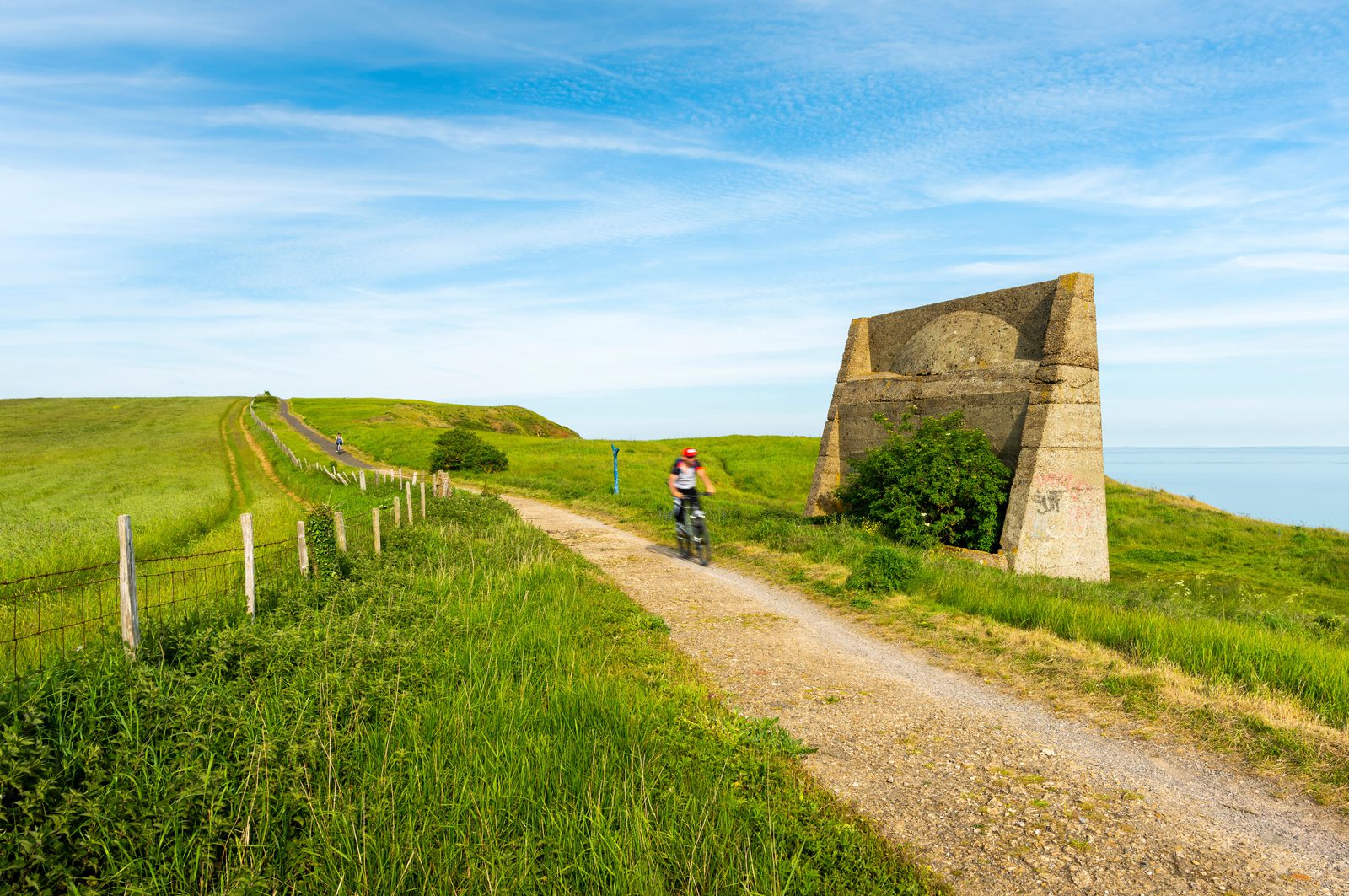 A person in a red helmet and backpack is riding a bicycle on a dirt path through a green grassy field. The path winds past an old, large concrete structure on the right, with a fence and more grassy hills in the distance under a blue sky with wispy clouds.