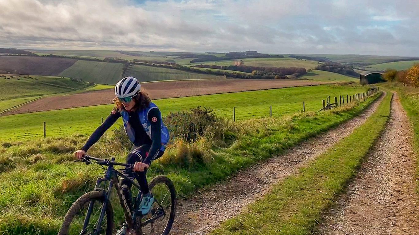 A person wearing a cycling helmet, sunglasses, and a blue jacket is riding a bicycle on a gravel path through green rolling hills under a cloudy sky. Fenced fields and distant woods can be seen in the background.