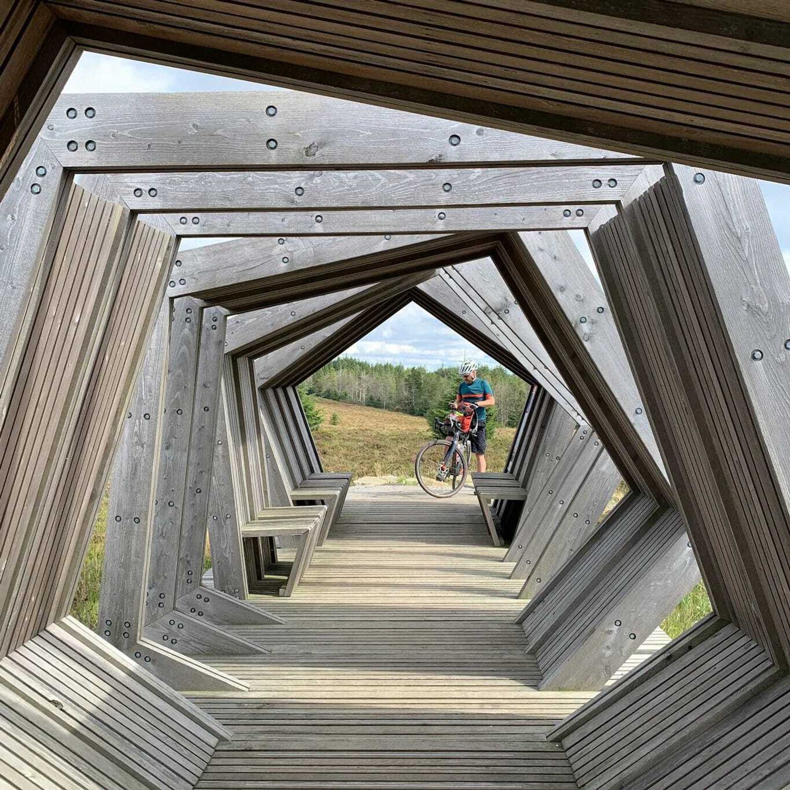 A cyclist pauses on a unique wooden bridge with geometric, angular structures forming a tunnel-like pathway. The bridge is set in a natural landscape with trees and grassy fields in the background.