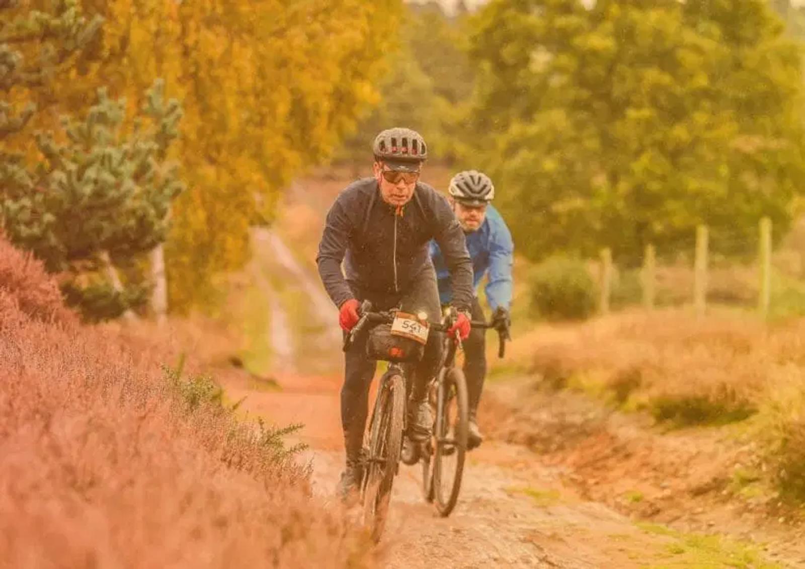 Two cyclists ride on a dirt path surrounded by autumn foliage. The lead cyclist wears a dark jacket and a helmet, while the second cyclist follows closely behind in a blue jacket. The scene is set in a countryside landscape with vibrant fall colors.