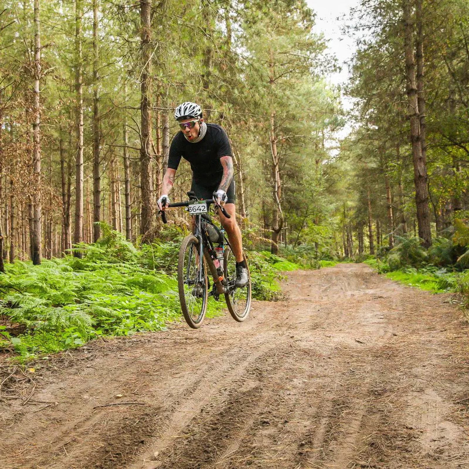 A cyclist wearing a helmet and sunglasses rides a bike on a dirt trail through a forest. The path is surrounded by lush green ferns and tall trees under a clear sky. The cyclist appears focused and determined.