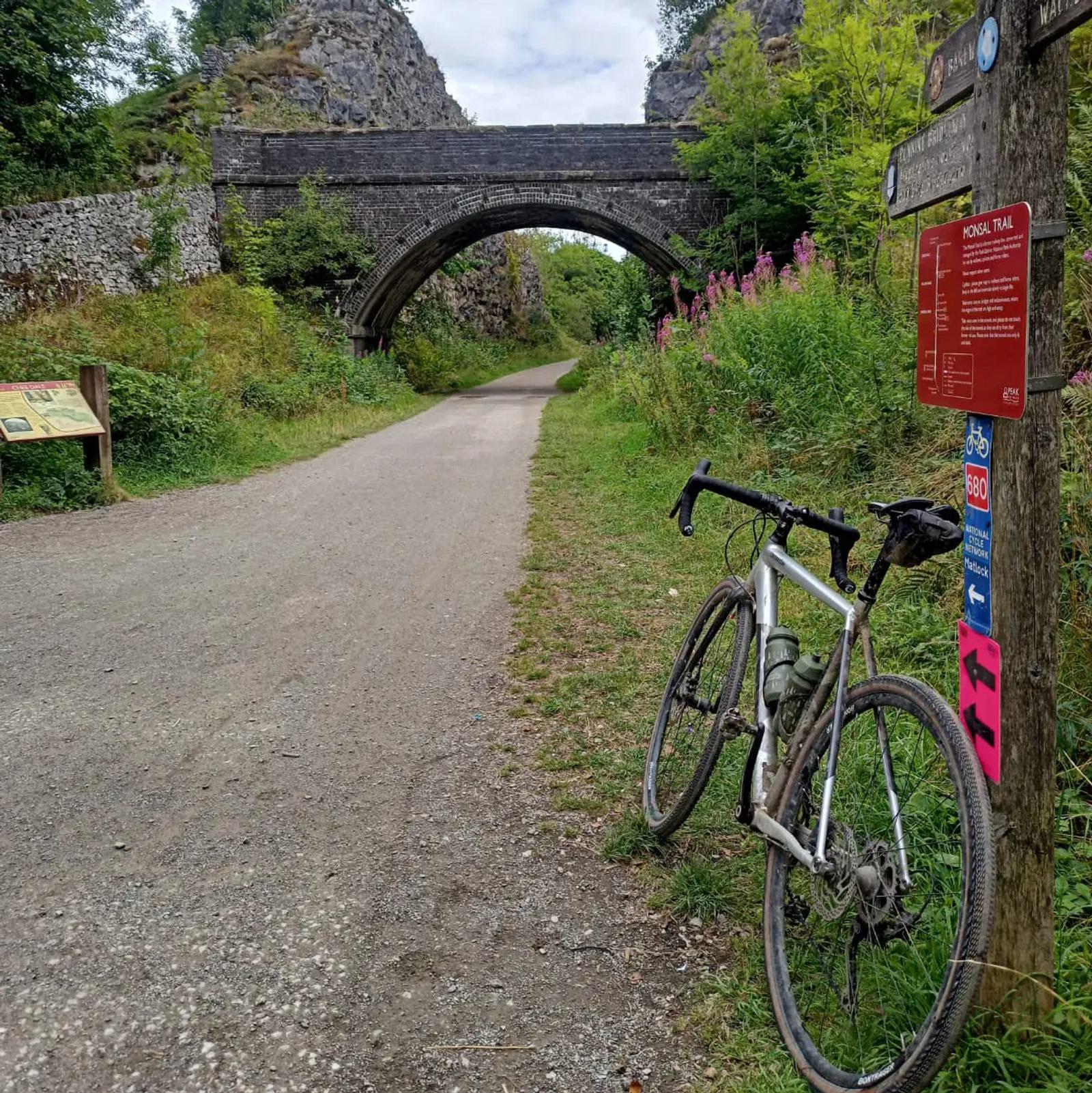A gravel bike is leaned against a signpost on a path leading to a stone bridge in a lush, green setting. Signs with information and direction arrows are visible. The path is surrounded by greenery and rocky hillsides.