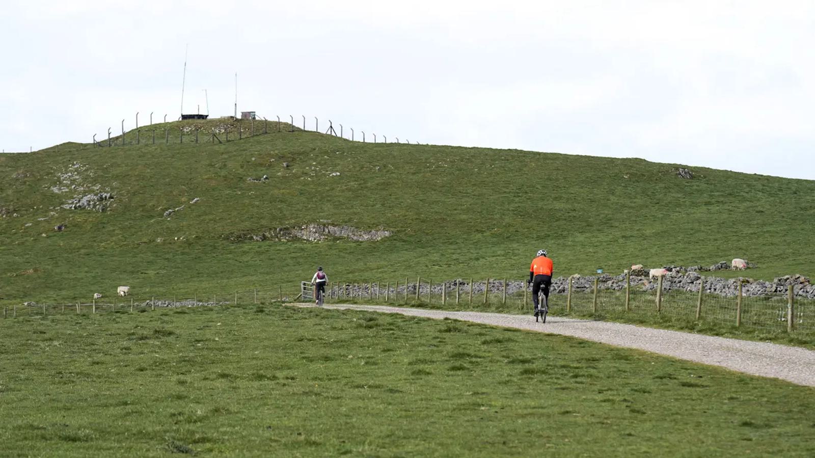 Two cyclists ride on a narrow gravel path through a vast, green landscape with rolling hills. One cyclist wears an orange top while the other wears a pink top. A small hill with a fenced structure is visible in the distance under a cloudy sky.