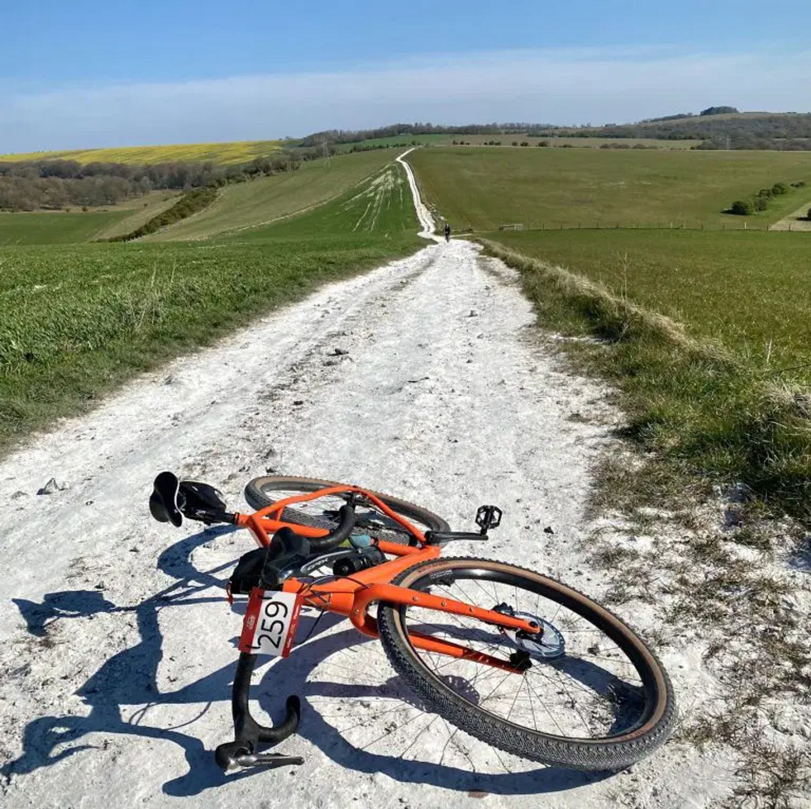 An orange bicycle lies on a white dirt path, numbered 259. The path stretches into the distance through green fields under a clear blue sky.
