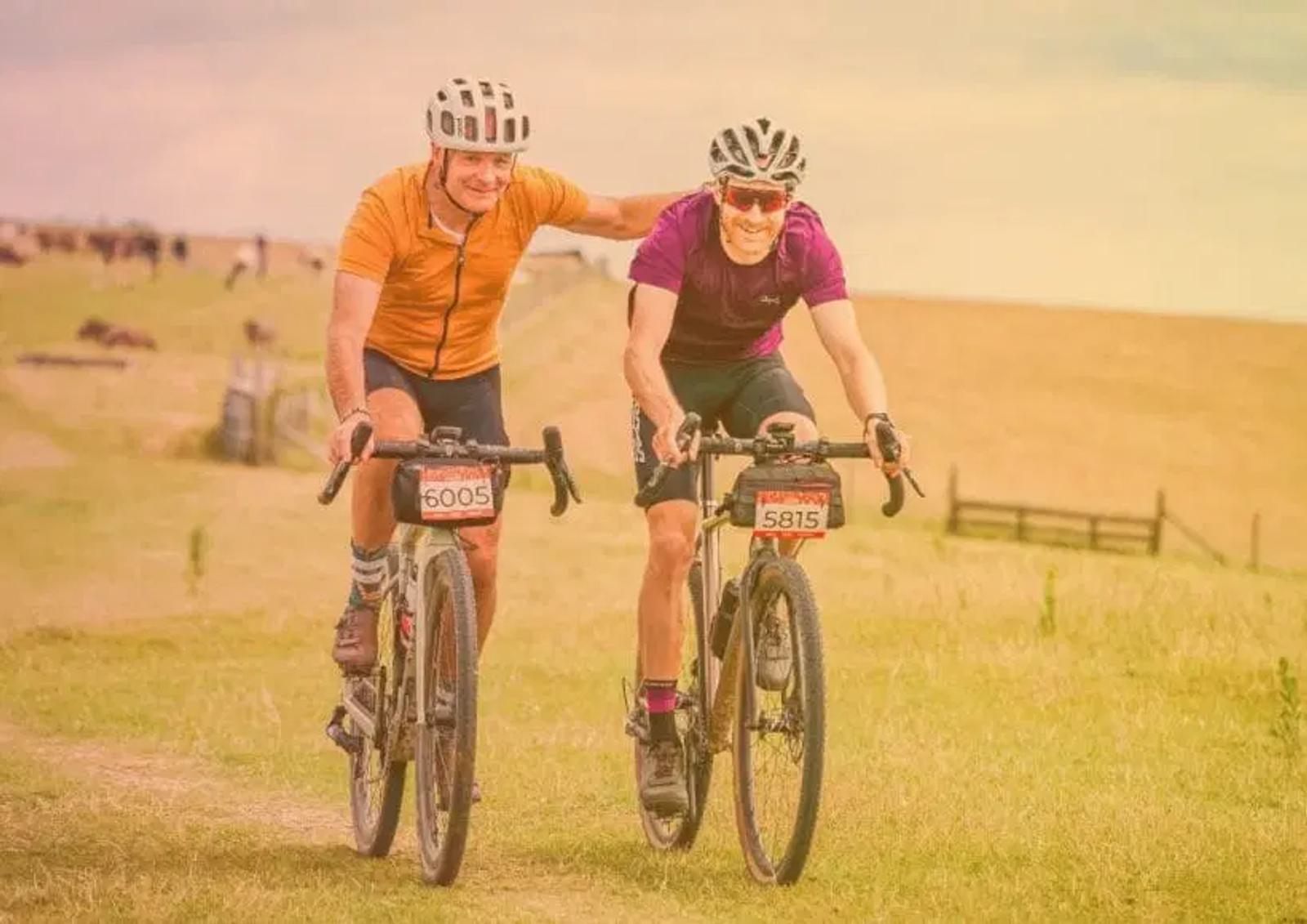 Two cyclists in helmets and jerseys ride on a grassy path, sharing a moment of camaraderie. The rider on the left wears an orange jersey and the one on the right wears a purple jersey. The background features rolling fields under a cloudy sky.