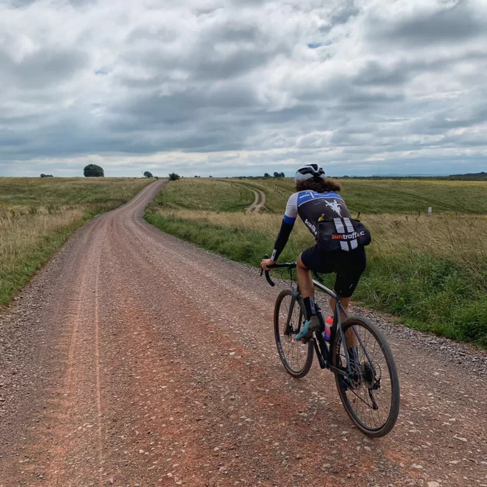 A cyclist rides along a long, winding gravel road through a grassy landscape under a cloudy sky. They wear a helmet and cycling gear, and the road stretches into the distance, flanked by fields.
