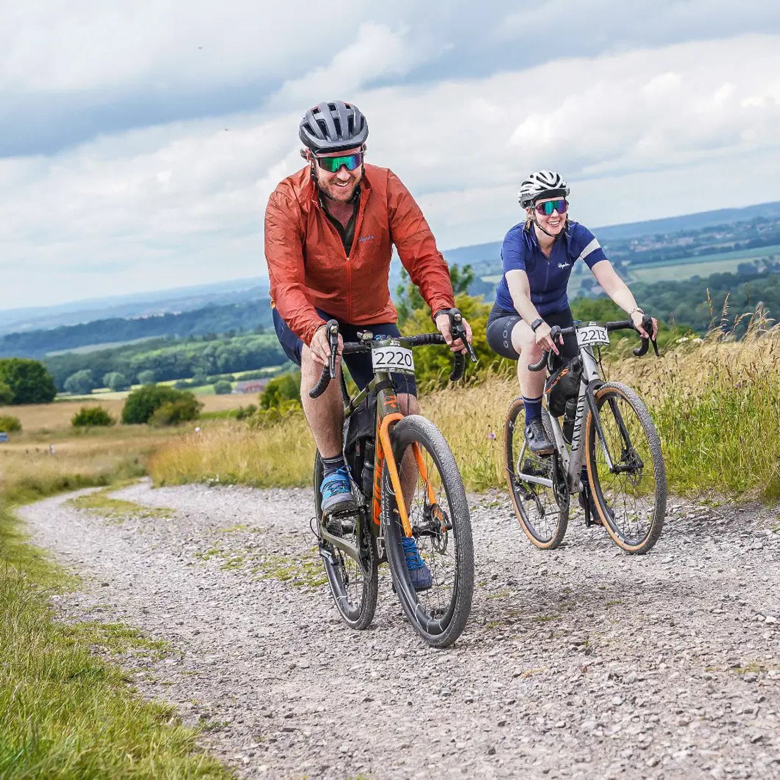 Two cyclists riding on a gravel path through a scenic landscape. The front cyclist wears a red jacket and the rear cyclist is in a blue outfit. Both have helmets and sunglasses, with numbered race bibs. Rolling fields are visible under a cloudy sky.