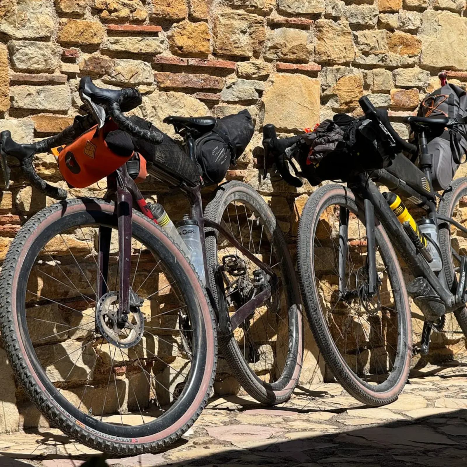 Three bicycles equipped with bags are parked against a rustic stone wall. The bikes have large tires and are set up for a cycling adventure, suggesting a focus on touring or bikepacking. The sunlight casts shadows on the cobblestone ground.