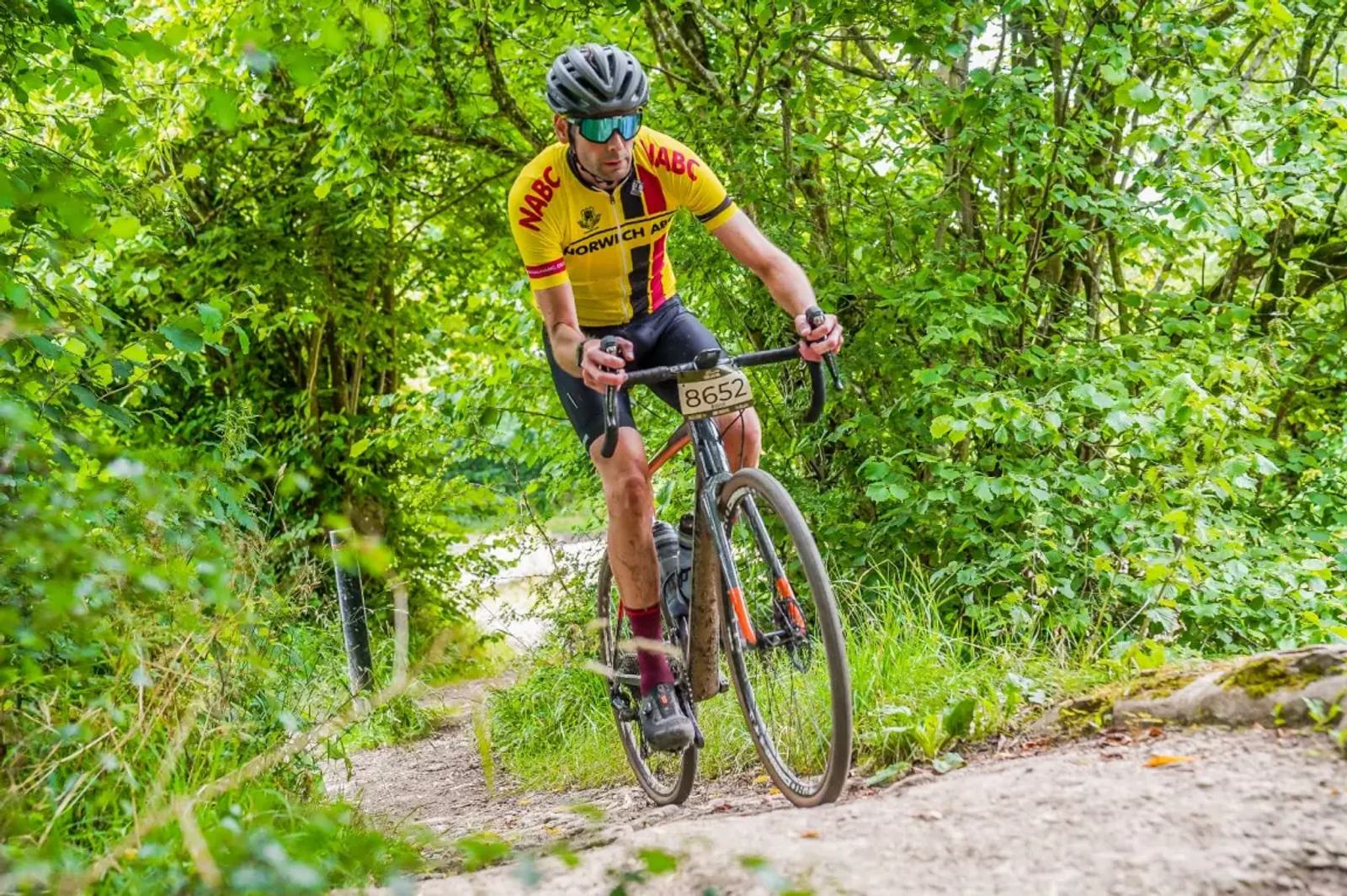 A cyclist wearing a yellow and red jersey and helmet rides a gravel bike on a forest trail. The path is surrounded by lush green foliage. The bike has a number tag, and the rider appears focused on the route ahead.