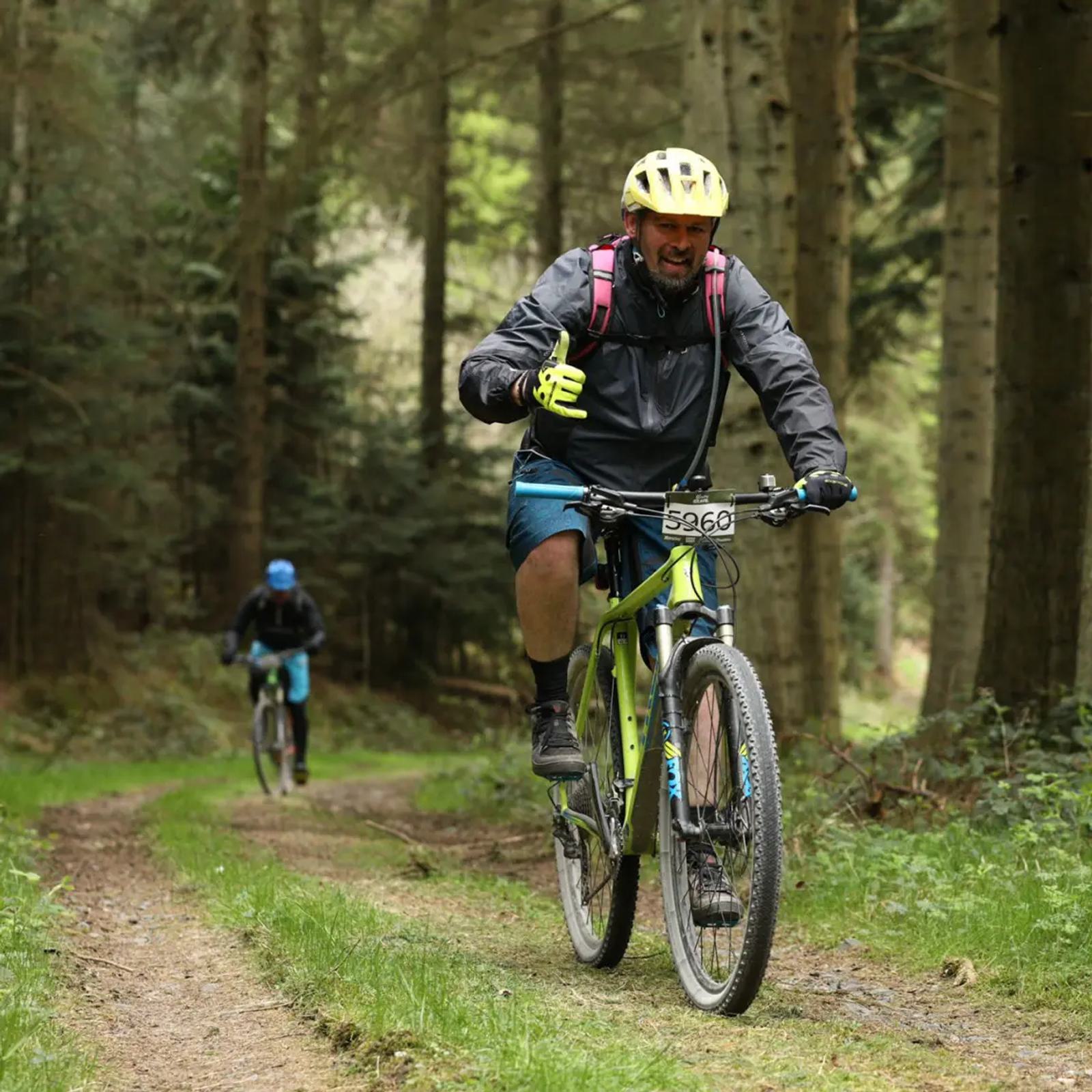 Two cyclists ride mountain bikes on a forest trail. The foreground rider wears a yellow helmet, black jacket, and blue shorts, smiling with a thumbs-up. A second cyclist follows in the background, surrounded by tall trees and greenery.