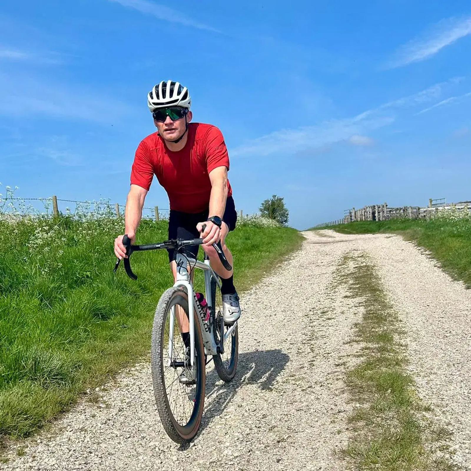 A person wearing a red shirt, black shorts, and a helmet rides a bicycle on a gravel path surrounded by green grass under a clear blue sky.