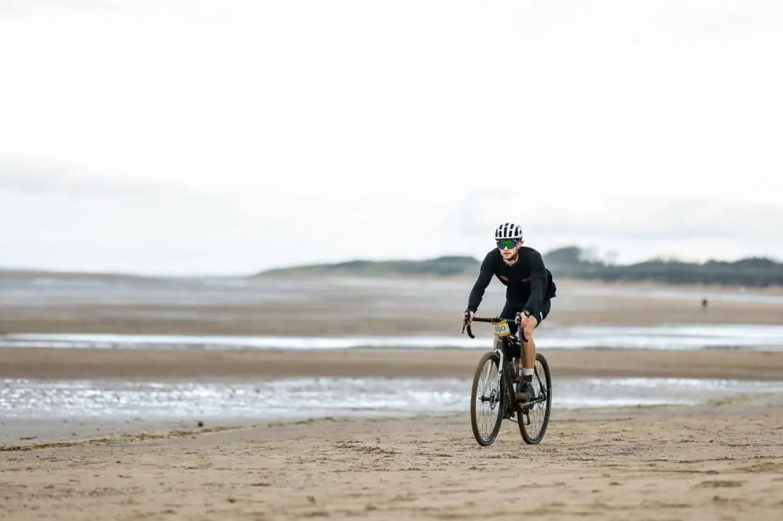 Cyclist with a striped helmet rides on a sandy beach, with a vast expanse of wet sand and ocean in the background. The sky is overcast, creating a serene and tranquil atmosphere.