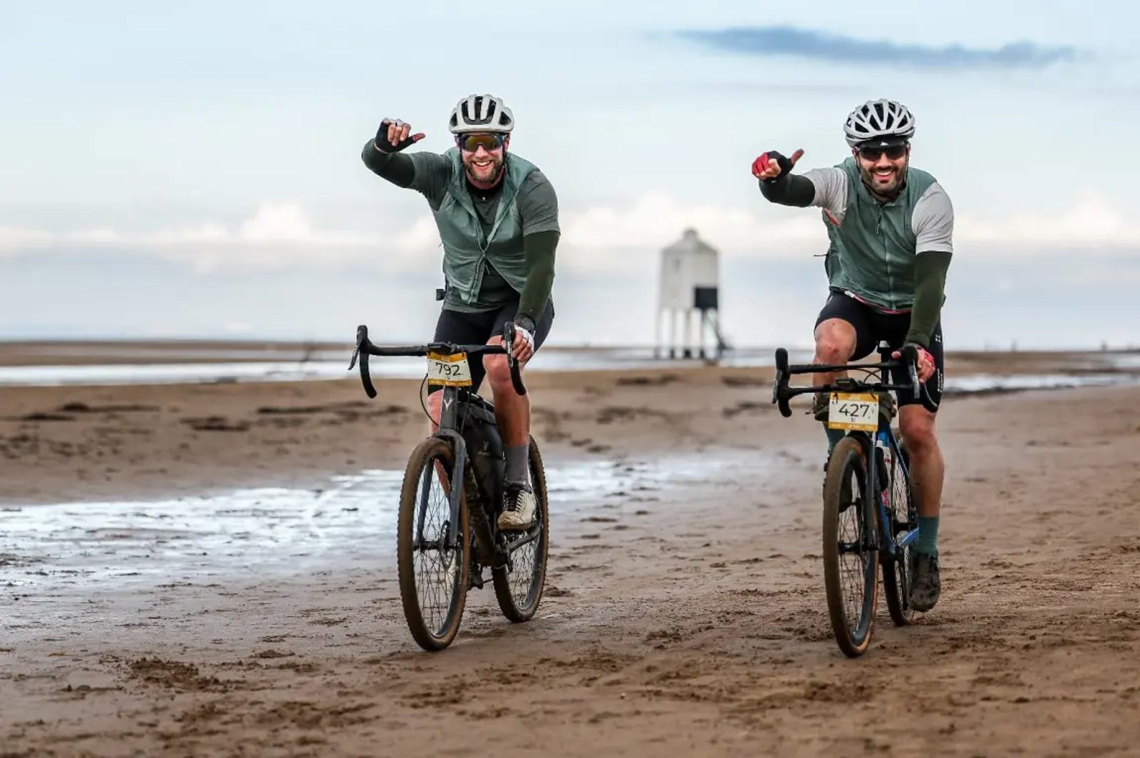Two cyclists ride along a beach, smiling and giving thumbs-up. They wear helmets, sunglasses, and numbered race tags. The background shows a lighthouse on a cloudy day.