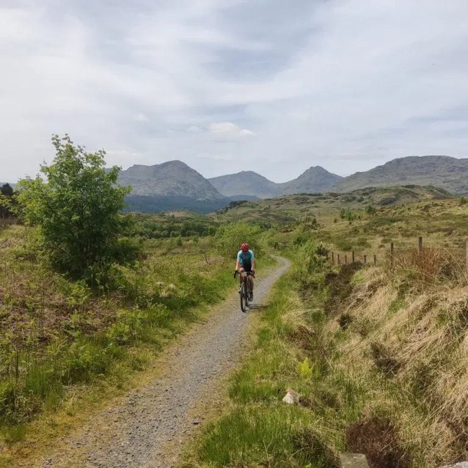A cyclist wearing a red helmet and turquoise shirt rides on a gravel path through a grassy landscape with distant mountains under a partly cloudy sky.