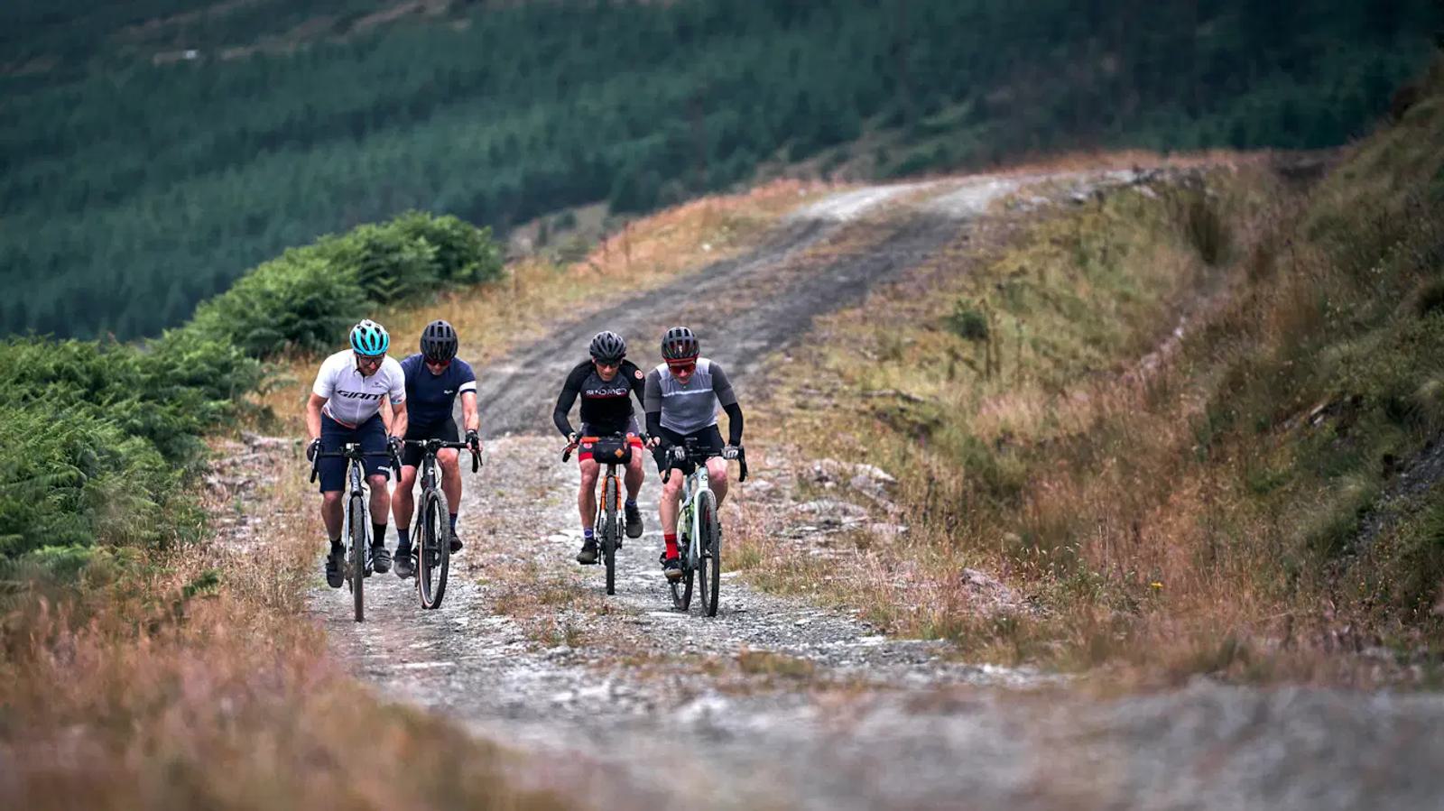 Four cyclists riding on a gravel path through a lush, hilly landscape. They are wearing helmets and casual cycling attire. Greenery lines the path, and a dense forest is visible in the background under an overcast sky.