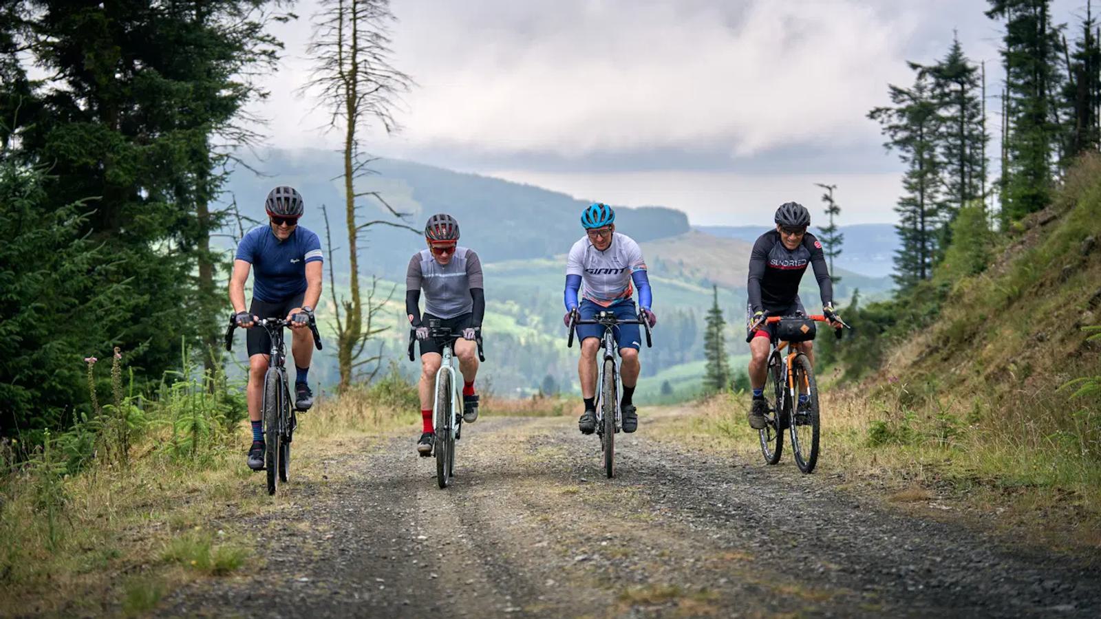 Four cyclists ride on a gravel path through a forested area, with hills and cloudy skies in the background. They wear helmets and cycling gear, appearing focused on their ride.