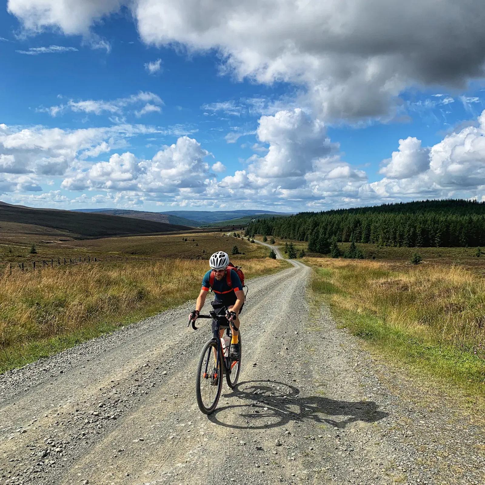 A cyclist rides on a gravel road through a scenic countryside with grassy fields and a forest in the background. The sky is partly cloudy with patches of blue.