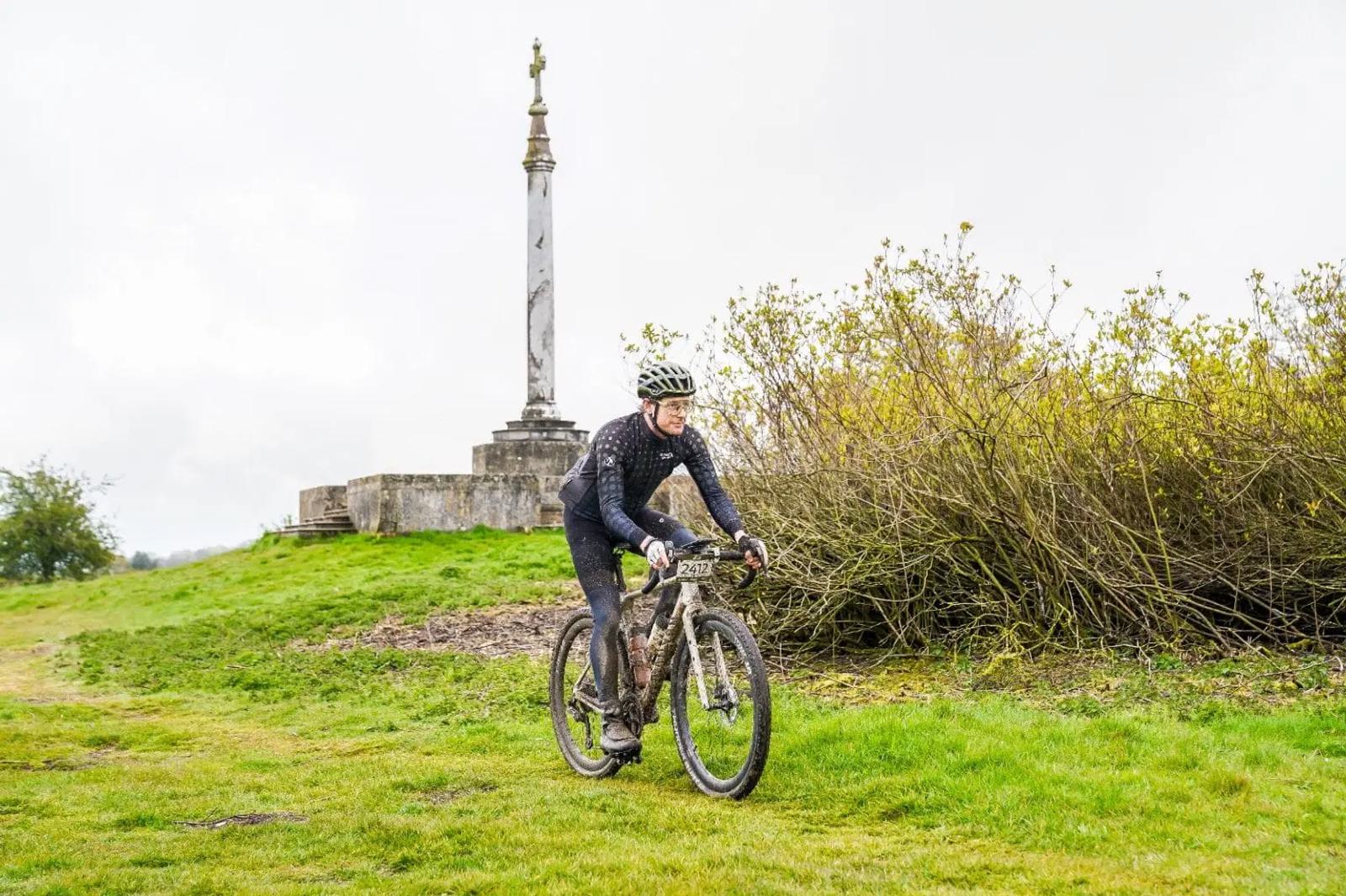 A cyclist navigates a grassy path on a muddy mountain bike, wearing a helmet and sports gear. In the background, there's a tall stone monument with a statue on top, surrounded by bushes under a cloudy sky.
