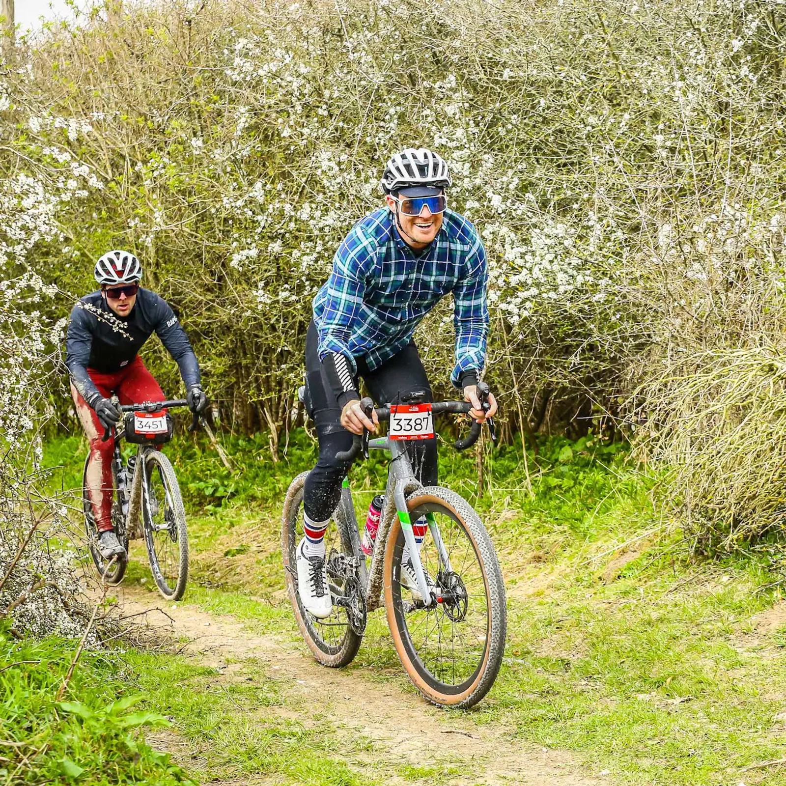 Two cyclists ride along a grassy trail surrounded by blooming bushes. The lead cyclist wears a checkered shirt and helmet, while the second cyclist follows closely behind in red pants. Both are focused and enjoying the outdoor ride.