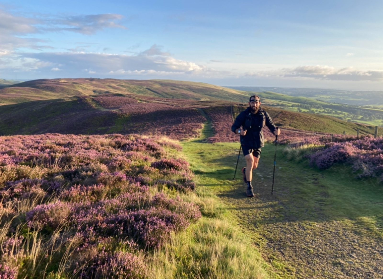 A person wearing hiking gear and sunglasses walks with trekking poles on a trail through a scenic landscape of rolling hills and purple heather under a blue sky with scattered clouds.