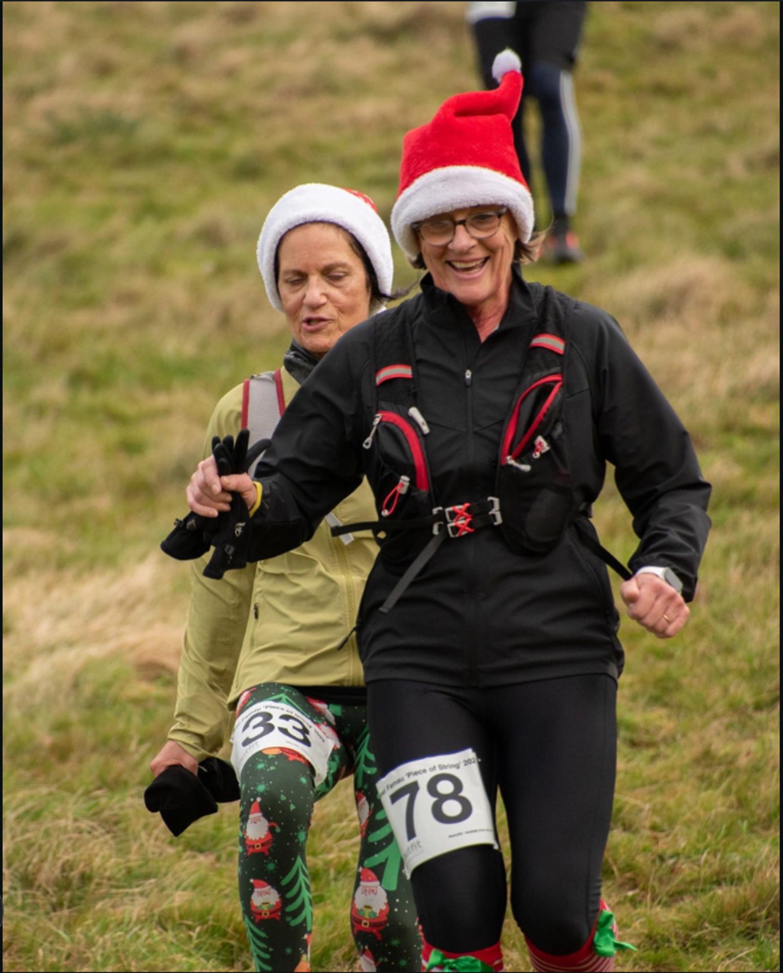 Two women running outdoors on a grassy field, both wearing festive Santa hats. The woman in front wears a black outfit and has a bib number 78. The woman behind wears light-colored clothing with a bib number 33 and holiday-themed leggings. Both appear joyful.
