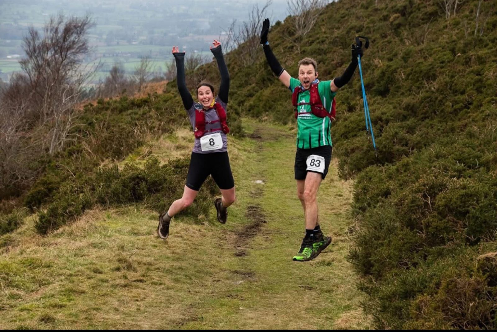 Two runners wearing race numbers jump joyfully on a grassy trail surrounded by hills. The woman on the left has her arms raised, and the man on the right holds walking poles and smiles. Bushes and distant fields are in the background.