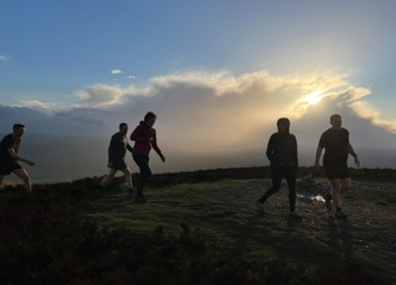 A group of five people in outdoor attire walk on a grassy hilltop with a stunning view of the sunset. The sky is partly cloudy, with the sun casting a warm glow over the landscape. The individuals are silhouetted against the bright backdrop.