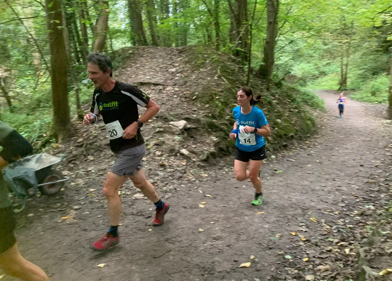 A man and a woman are running on a trail through a wooded area, wearing race bibs number 20 and 14, respectively. The ground is uneven with some fallen leaves. A person in the background follows behind them on the same path.