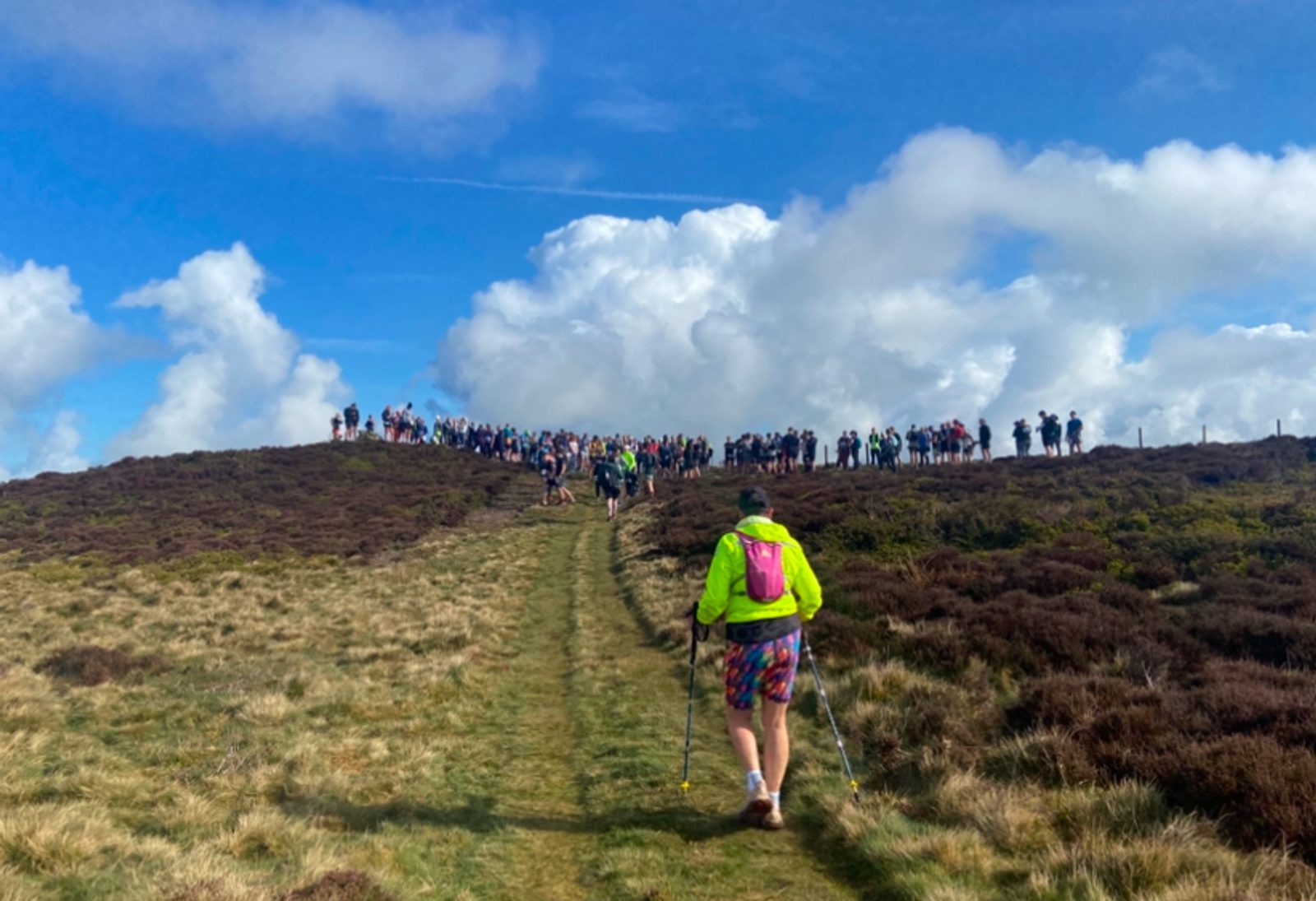 A person with hiking poles and a bright green jacket walks up a grassy trail towards a hilltop where a large group of people is gathered under a blue sky with scattered clouds.