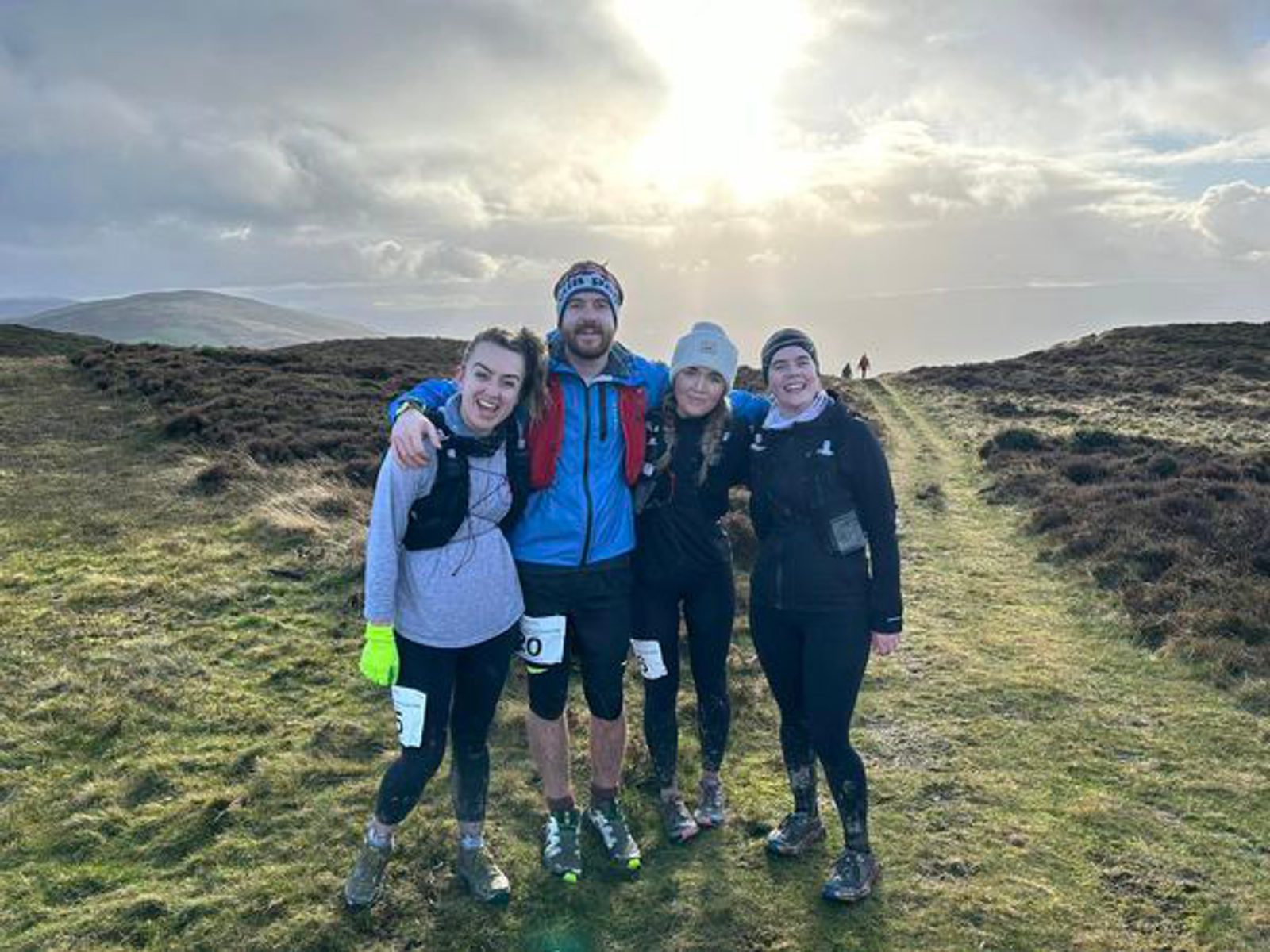 Four people pose together on a grassy hilltop during a hike or run. They are dressed in outdoor and athletic gear, including jackets, hats, and gloves. The sky is partly cloudy with the sun peeking through, casting a bright glow over the landscape behind them.