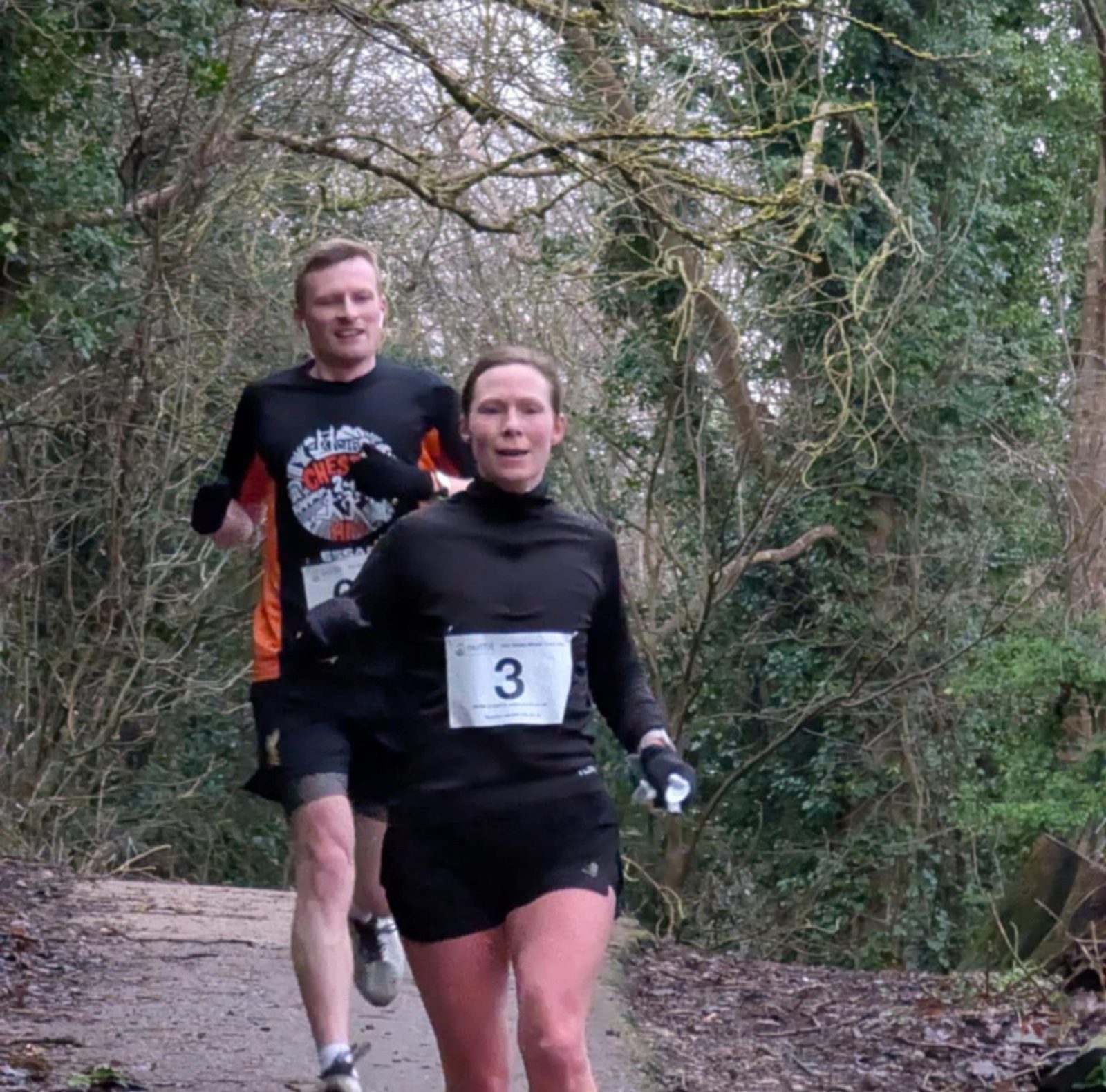 Two runners compete on a wooded trail. The woman in front wears a black outfit with bib number 3, and the man behind wears a black shirt with orange sleeves. Trees with bare branches line the path.