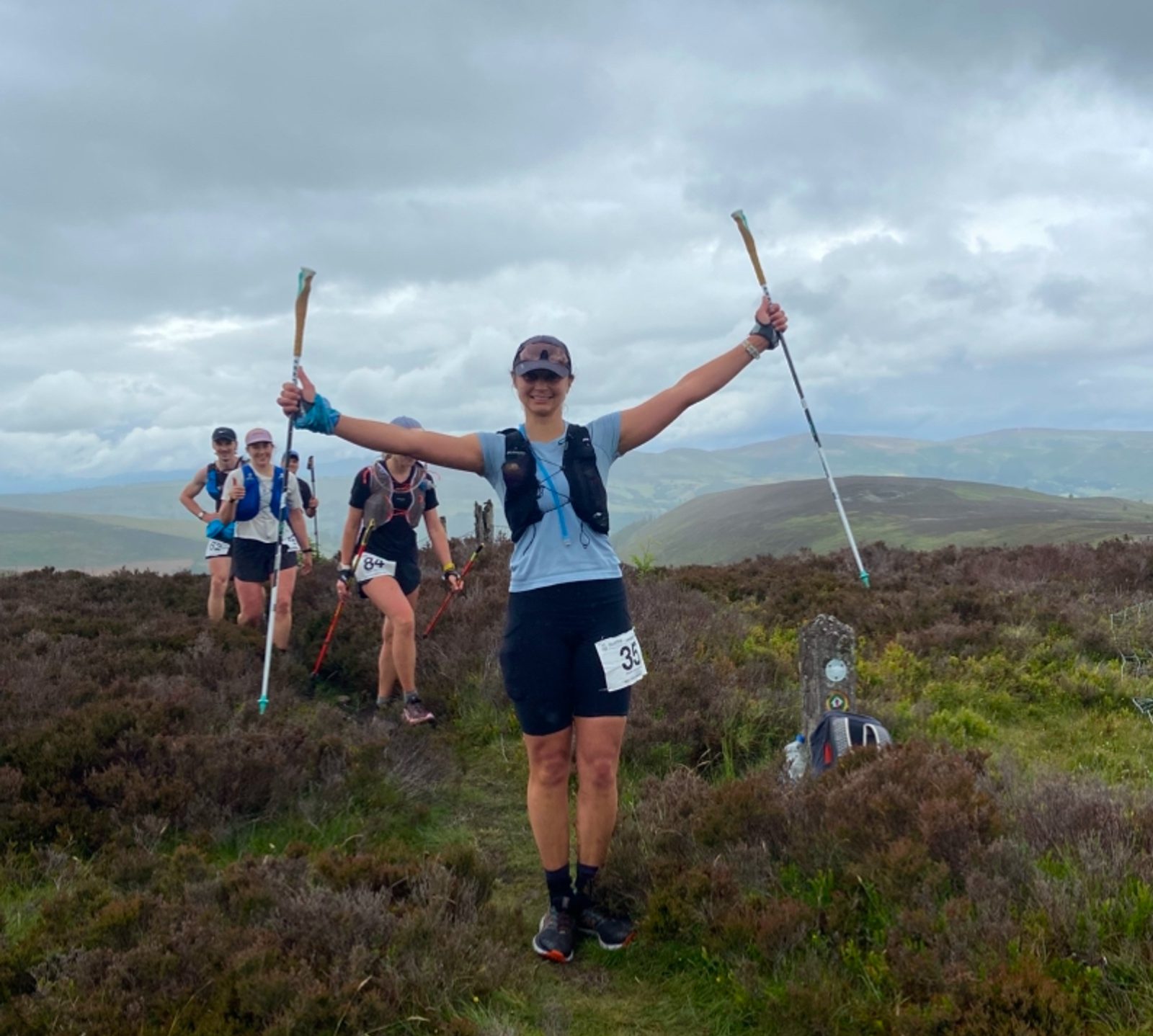 A group of trail runners, each holding poles, move along a heather-covered path on a hilltop. One runner in front smiles with arms raised in triumph. Rolling hills and cloudy skies are in the background.