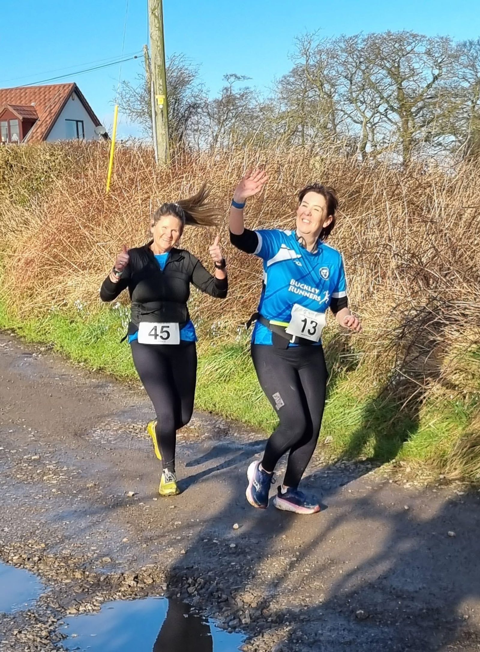 Two women wearing race bibs run along a rural path, smiling and waving at the camera. The sun is shining, and they are surrounded by bushes and trees.