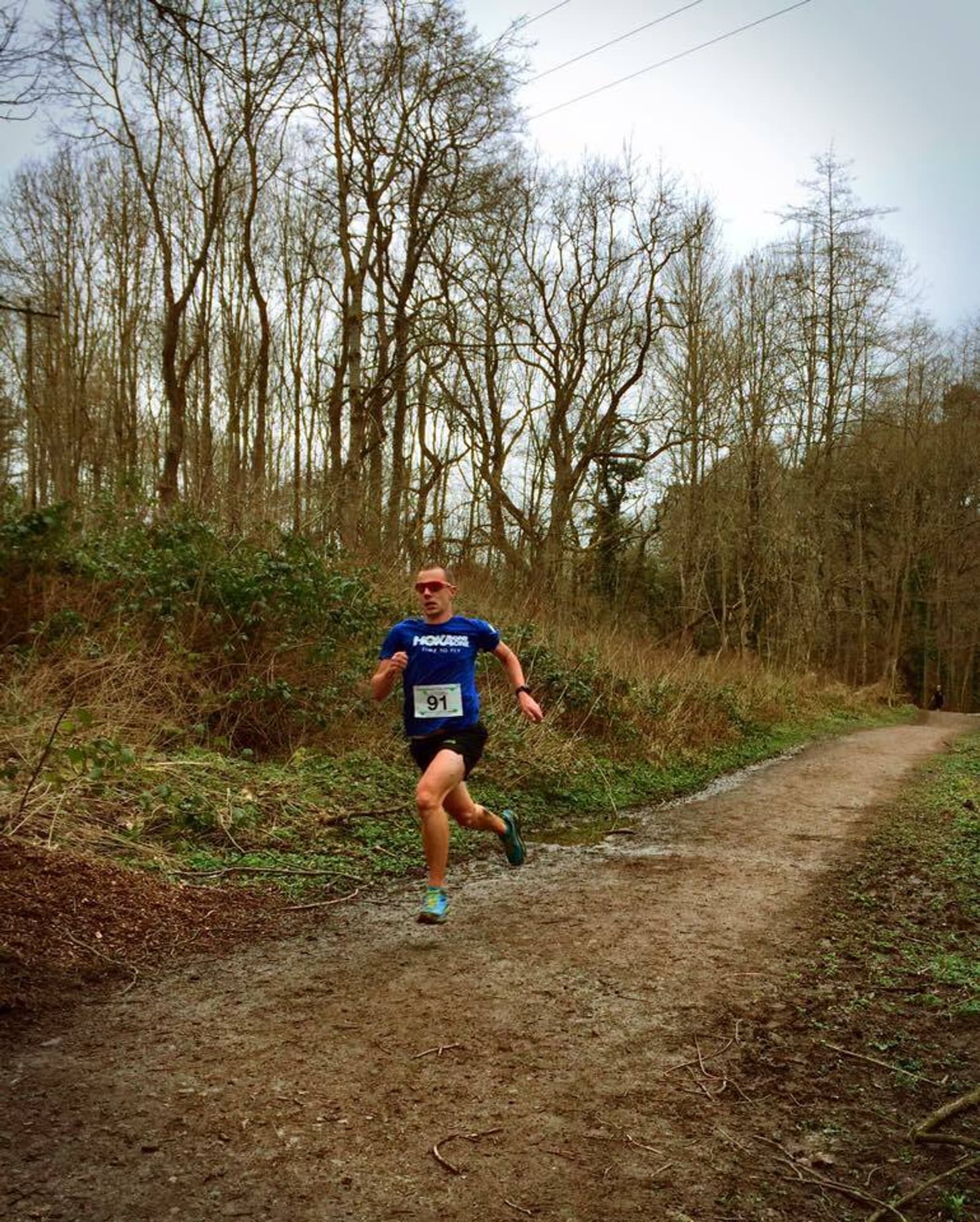 A runner wearing a blue shirt, black shorts, and teal shoes is participating in a race, running on a dirt path through a forested area. The runner is wearing a race bib with the number 91. Leafless trees and dense undergrowth are visible in the background.