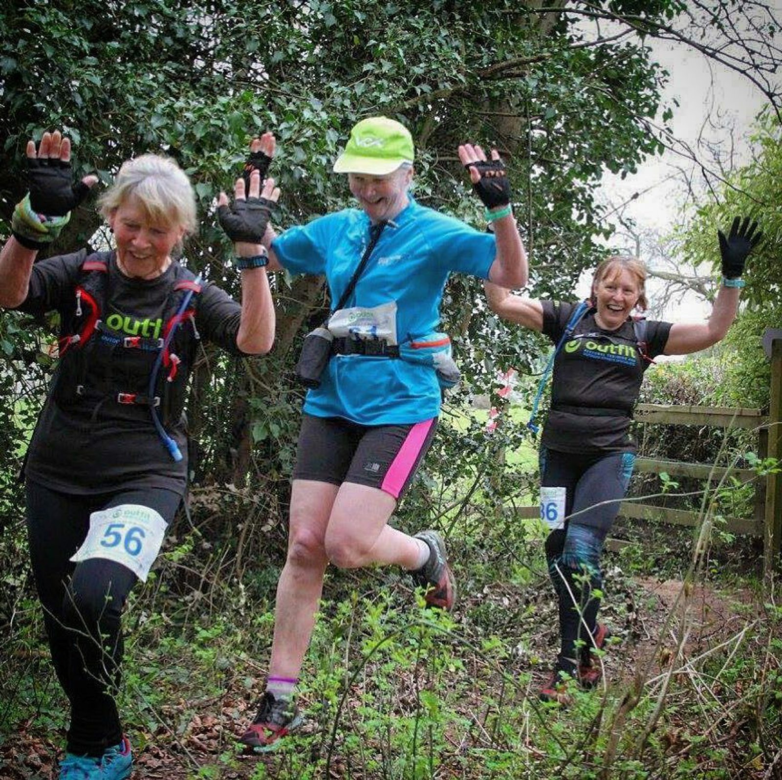 Three women in athletic gear are running through a wooded trail, laughing and raising their hands in a joyful gesture. They are wearing numbers on their outfits and carrying small backpacks, indicating participation in an organized outdoor event.