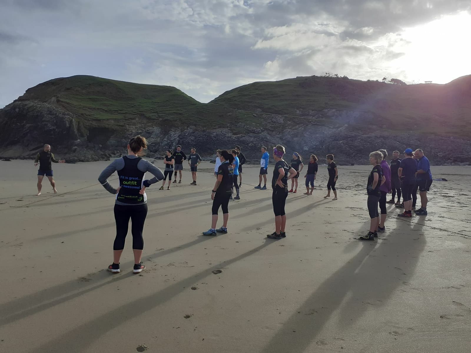 A group of people standing in a large circle on a beach during the daytime, with a rocky, green hill and cloudy sky in the background. Each person is dressed in athletic gear, and their long shadows stretch out on the sand.