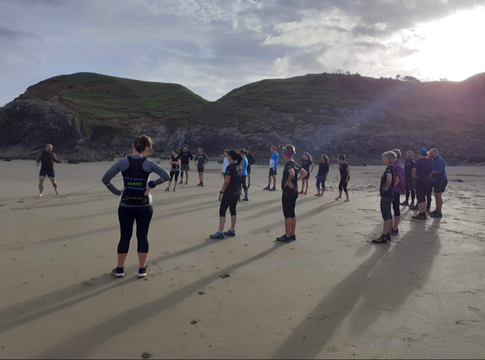 A group of people dressed in wetsuits stands spaced apart on a sandy beach beneath a cloudy sky with sunlight breaking through. They face different directions, appearing to prepare for an activity, with rocky hills and cliffs in the background.