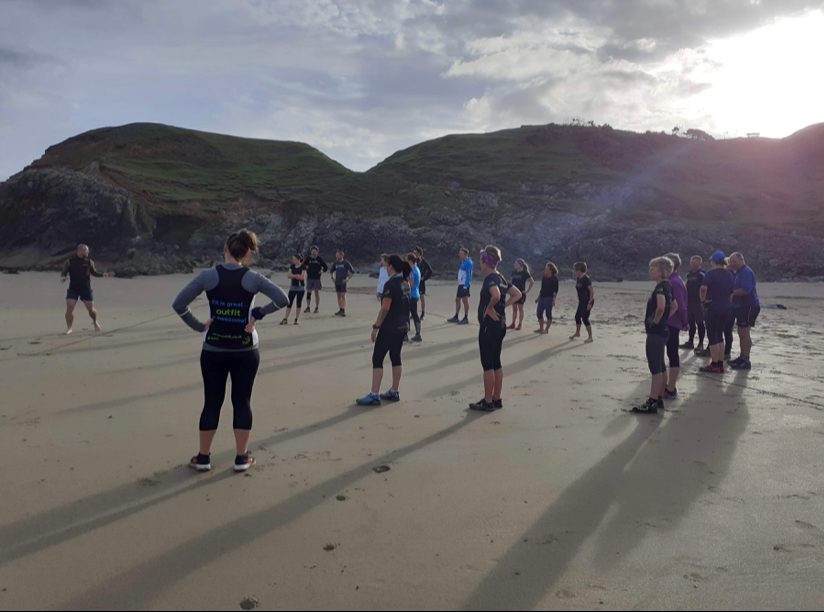 A group of people dressed in wetsuits stands spaced apart on a sandy beach beneath a cloudy sky with sunlight breaking through. They face different directions, appearing to prepare for an activity, with rocky hills and cliffs in the background.