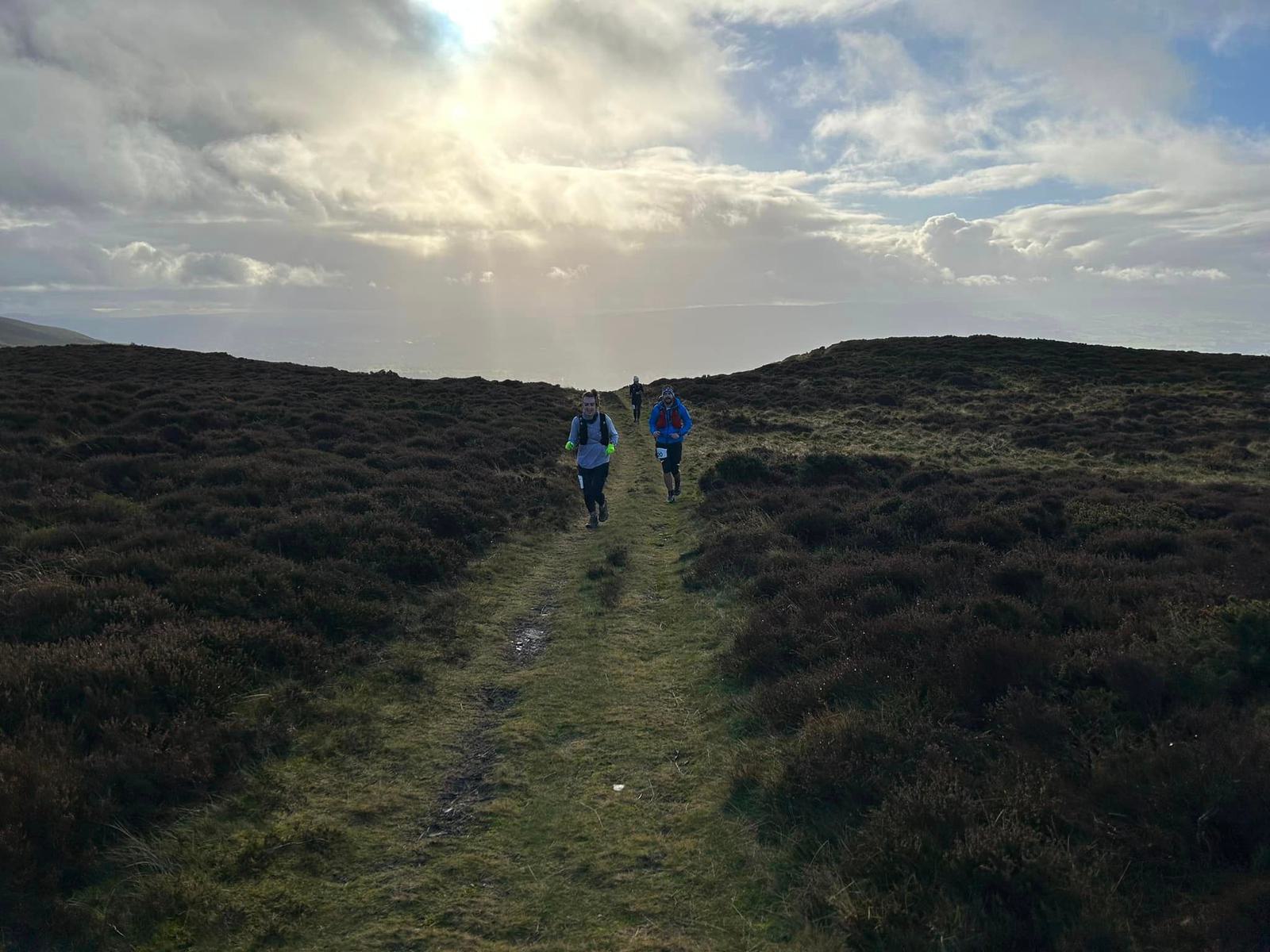 Two people are running on a grassy path through a hilly, moor-like landscape under a partly cloudy sky with the sun peeking through. The trail is flanked by shrubs and low vegetation, and the horizon is dimly visible in the distance.