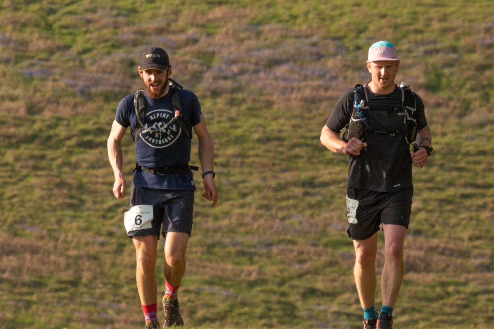 Two men are running on a grassy path with gentle slopes in the background. Both wear black shorts, t-shirts, and caps. Each has a small runner's pack strapped on their backs, and visible bib numbers. Their expressions suggest concentration on their running.