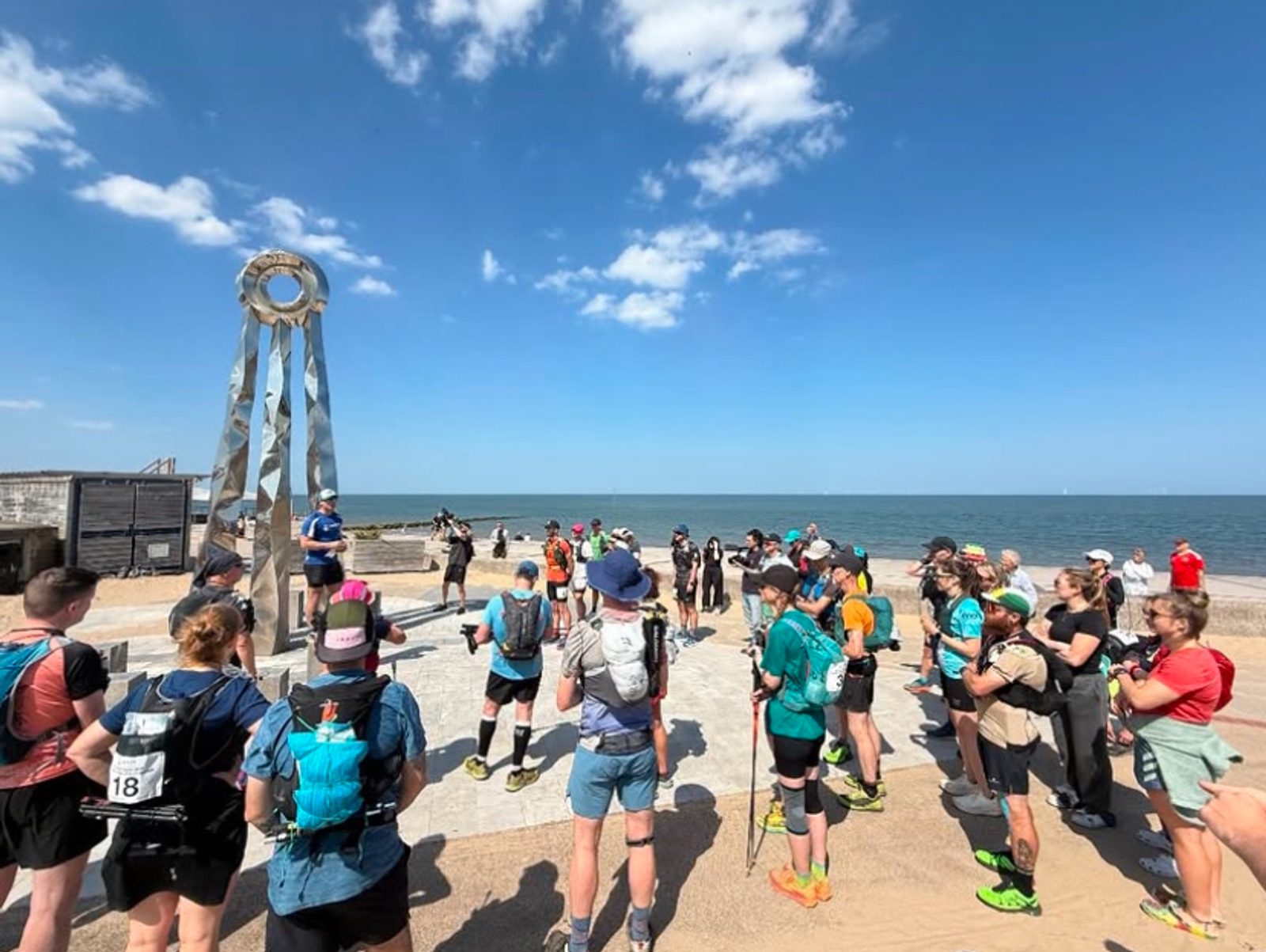 A group of runners with packs and numbered bibs gather near a modern metal sculpture by the beach, listening to instructions under a bright blue sky with a few clouds. The sea is visible in the background.