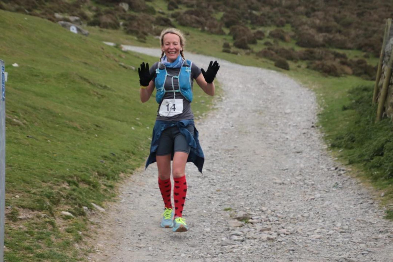 A person wearing a blue jacket, blue running vest, and red heart-patterned knee-high socks runs and smiles along a gravel path on a grassy, hilly terrain. The runner has black gloves and a race bib with the number 14.