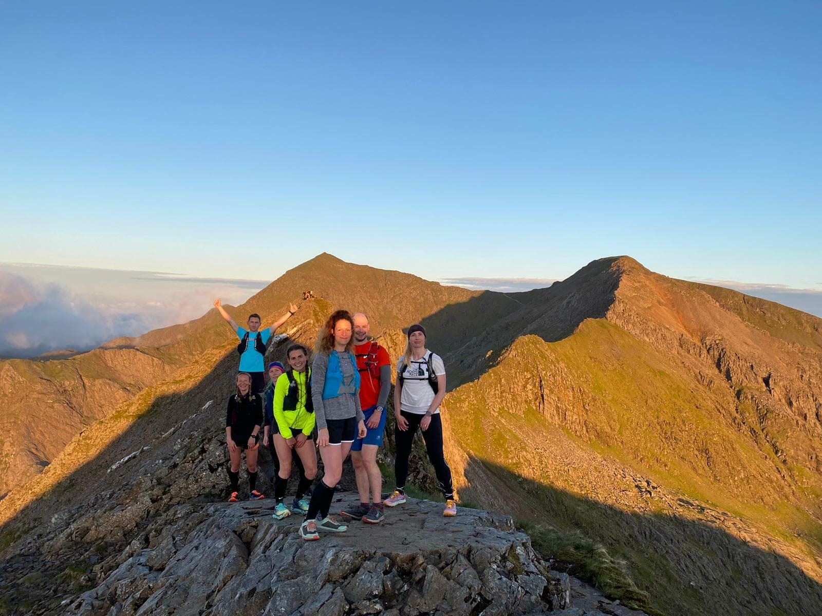 A group of six people standing on a rocky mountain ridge during sunset. They are dressed in hiking gear and appear happy, with one person raising their arms in excitement. The mountain peaks in the background are bathed in the warm golden light of the setting sun.