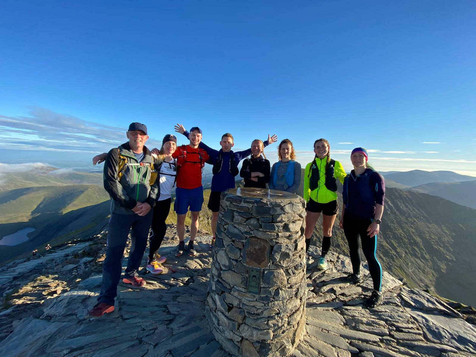 A group of eight people stand on a mountain summit by a stone marker under a clear blue sky. They are dressed in outdoor gear, smiling, and some have their arms raised in celebration. The surrounding mountains and valleys are bathed in sunlight.