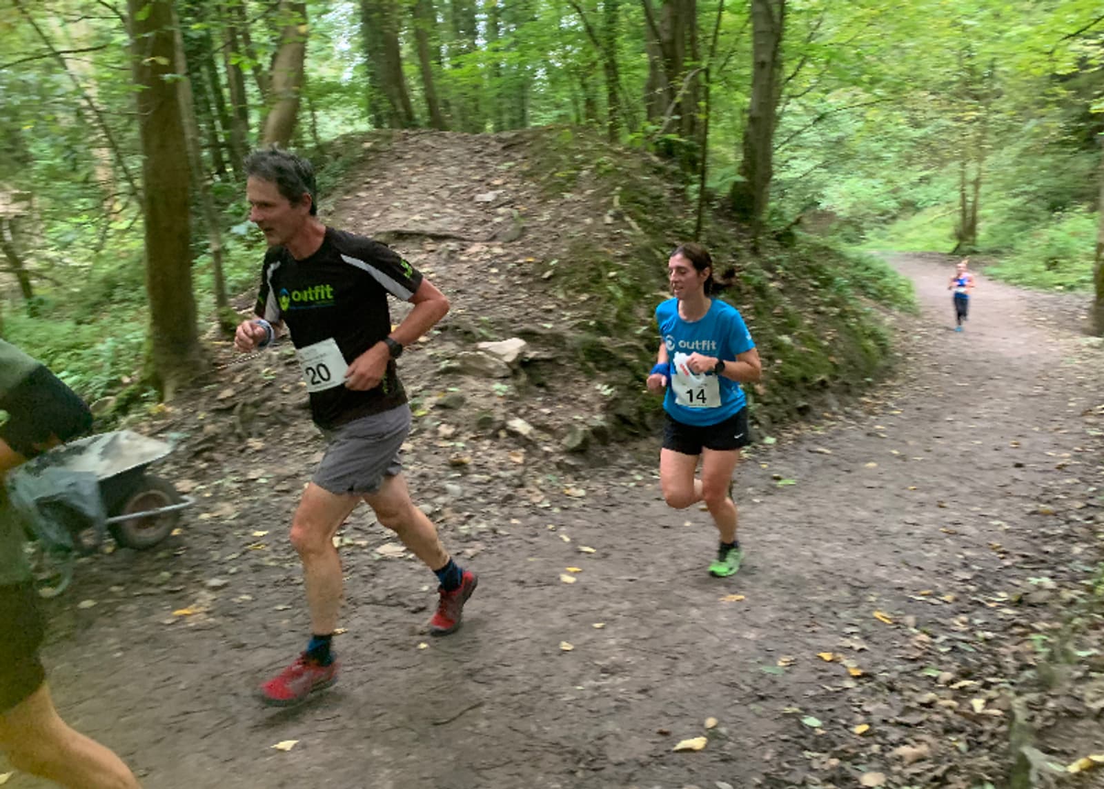 Two runners, both wearing numbered bibs (20 and 14), are running along a dirt trail in a forested area. They are surrounded by lush green trees. Additional runners can be seen further along the trail in the background. The ground is scattered with fallen leaves.