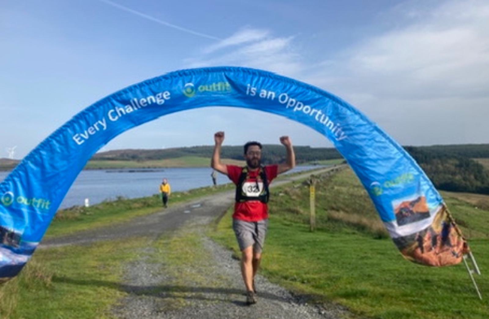 A man wearing a red shirt and a race bib crosses a finish line banner that reads "Every Challenge Is an Opportunity." He raises his arms in triumph. The path is surrounded by greenery and a body of water is visible in the background. The weather is clear.