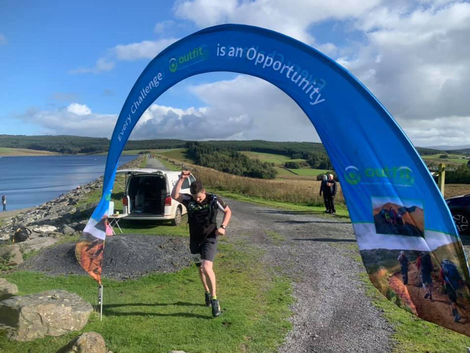 A runner wearing athletic gear sprints towards the finish line at an outdoor event. The finish line is marked by a blue arch with the words "Every Challenge is an Opportunity" printed on it. The background features a scenic landscape with a lake and rolling hills.