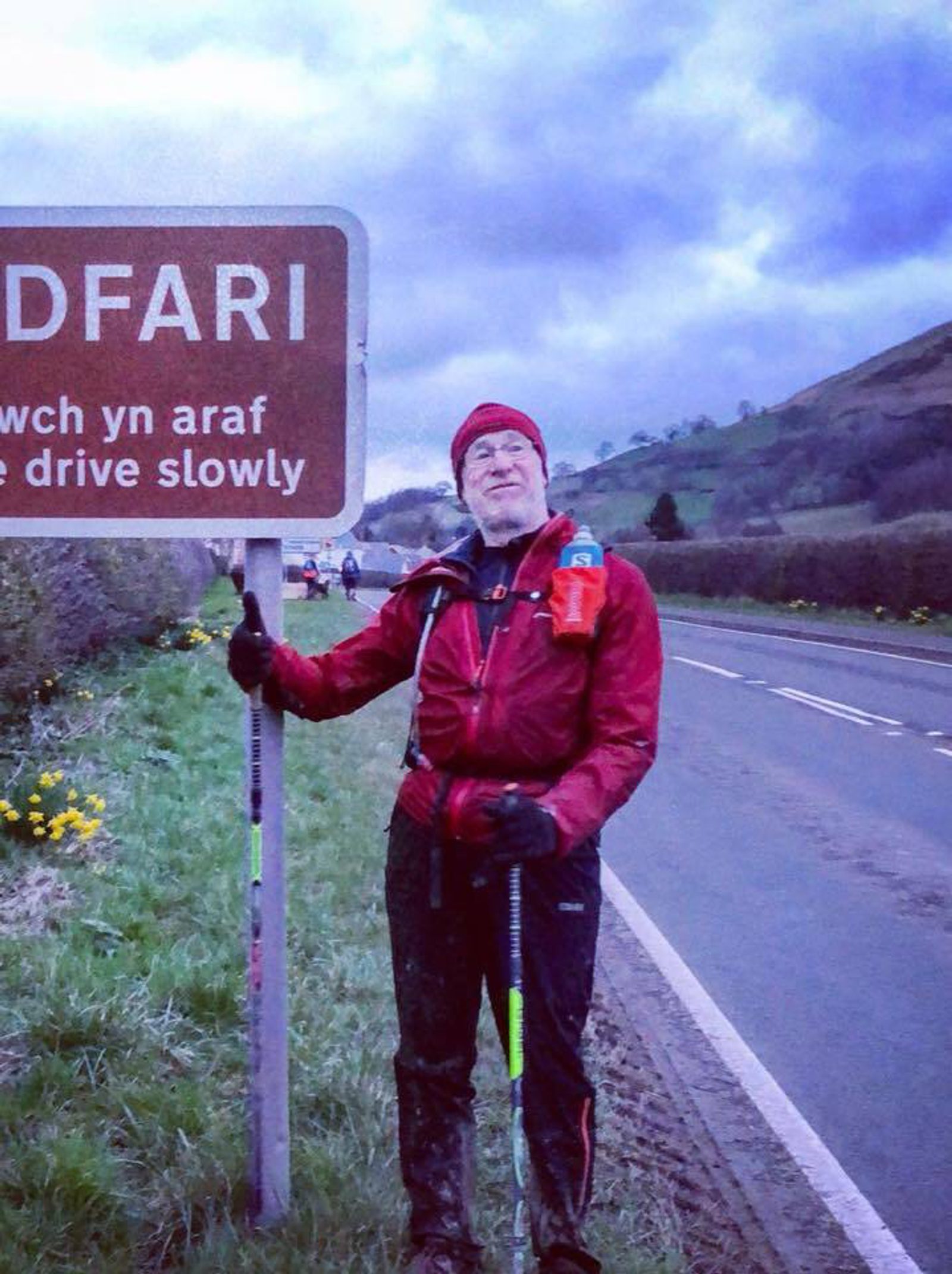 A person wearing a red jacket and hat stands by a roadside sign that reads "ARAF DWFARI Slow drive slowly." They hold onto a trekking pole and have a hydration backpack. The background features a winding road, grassy fields, and a cloudy sky.