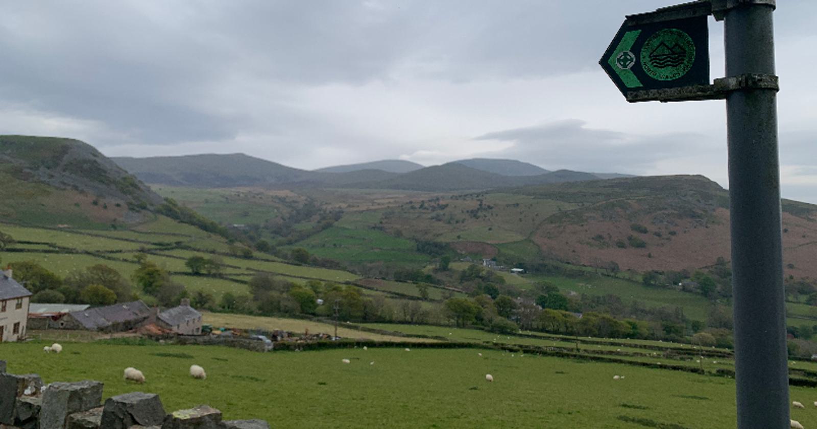 A scenic view of a lush valley with green fields and grazing sheep. Rolling hills and mountains are visible in the background under a cloudy sky. A wooden trail sign with a green marker is in the foreground, pointing to the right. Stone walls and houses are at the edge.